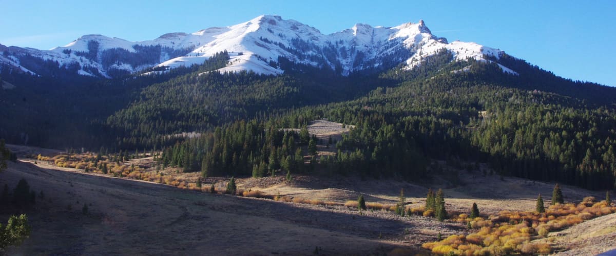 A mountain range with snow-capped peaks, a valley with autumn-colored foliage, and a clear blue sky.