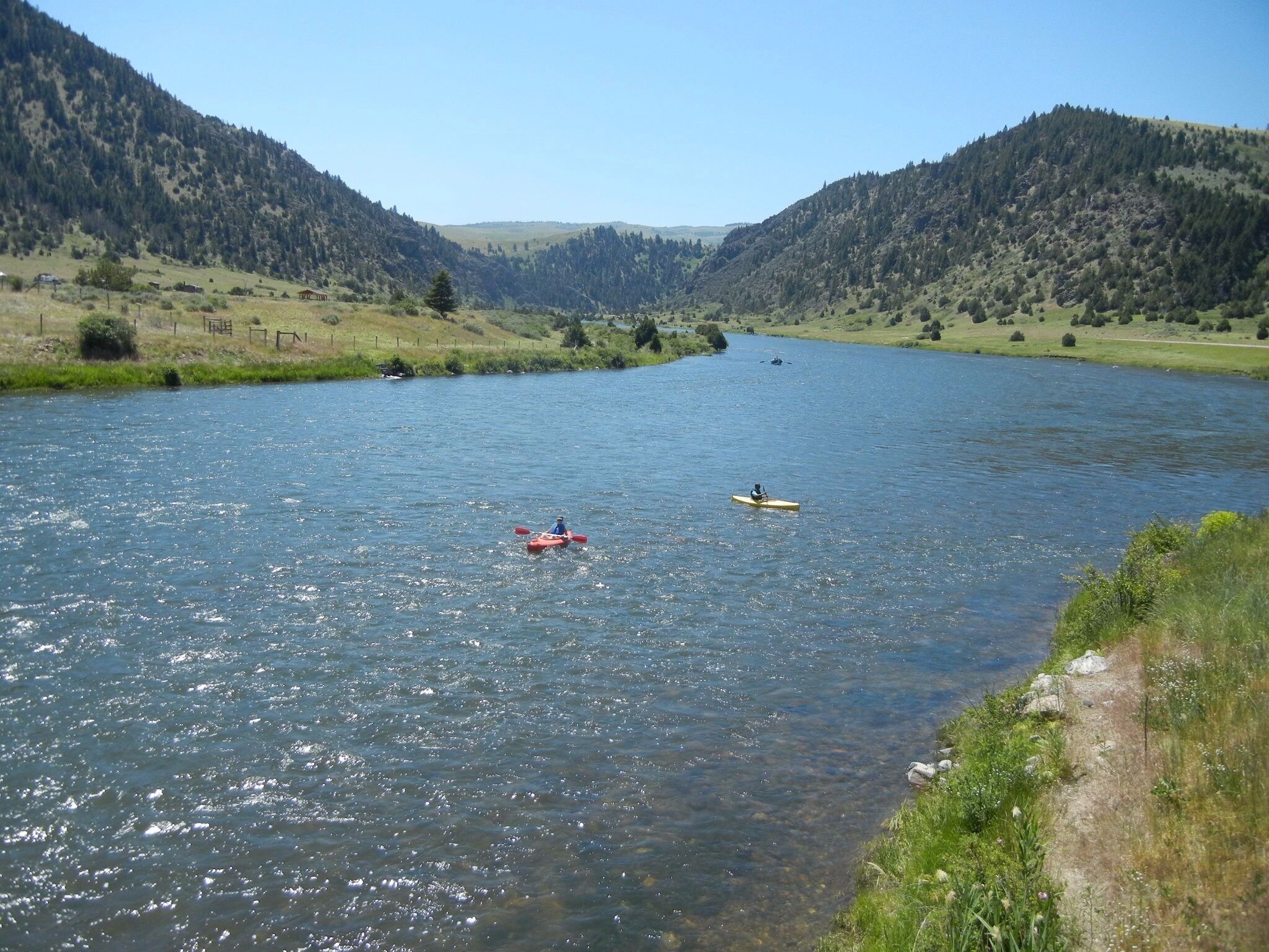 A river with kayakers, surrounded by green hills and a clear sky.