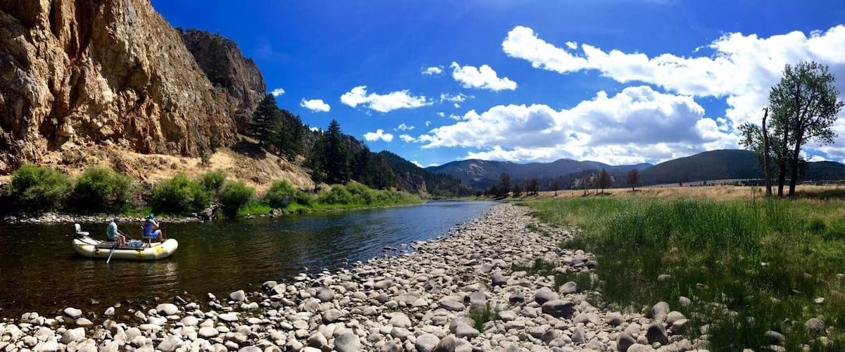 A river with a rocky shore, a raft with people, and mountains in the background.