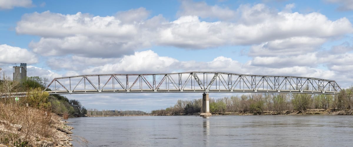 A steel truss bridge over a river with trees and a cloudy sky.
