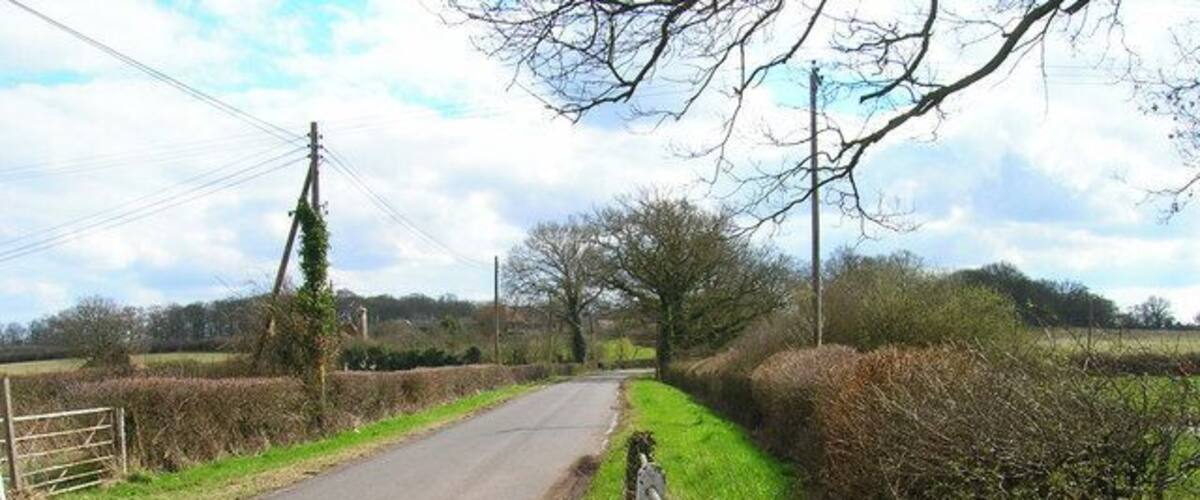 A narrow rural road flanked by hedgerows and a metal gate.