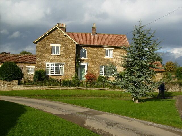 A stone house with a red-tiled roof, a green lawn, and a large evergreen tree.