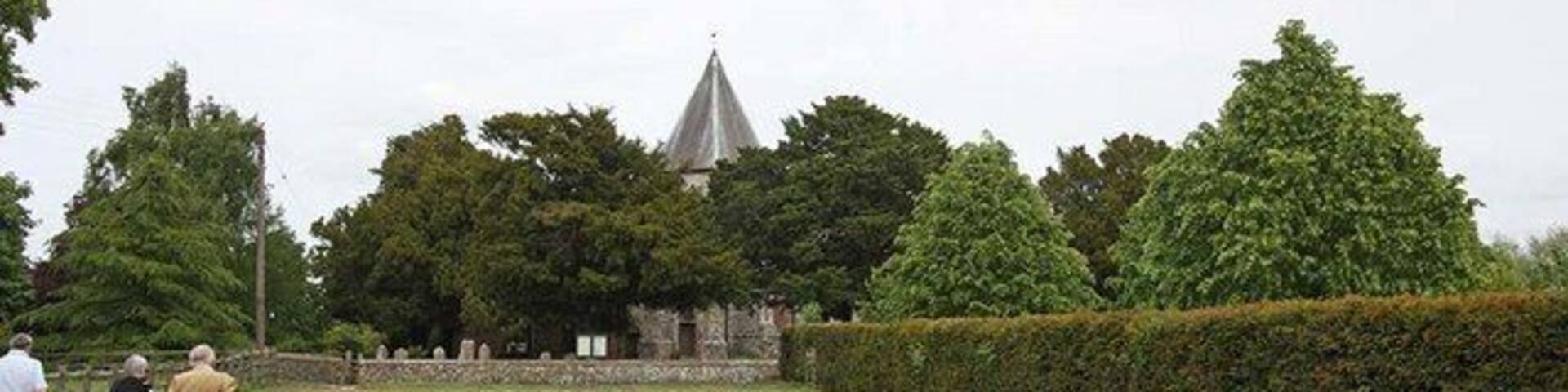 A cemetery with neatly trimmed hedges, a church with a pointed steeple, and a group of people walking.