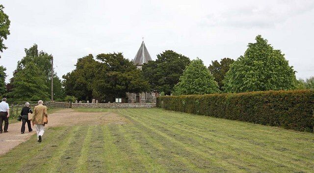 A cemetery with neatly trimmed hedges, a church with a pointed steeple, and a group of people walking.