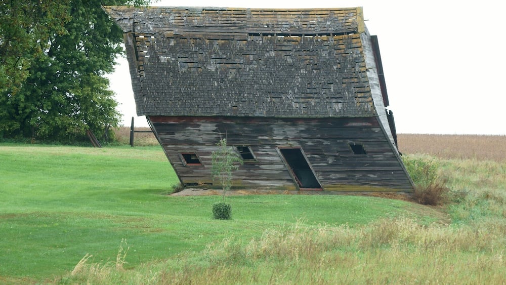 A tilted, dilapidated wooden structure in a grassy field with a tree in the background.