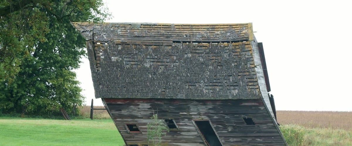 A tilted, dilapidated wooden structure in a grassy field with a tree in the background.