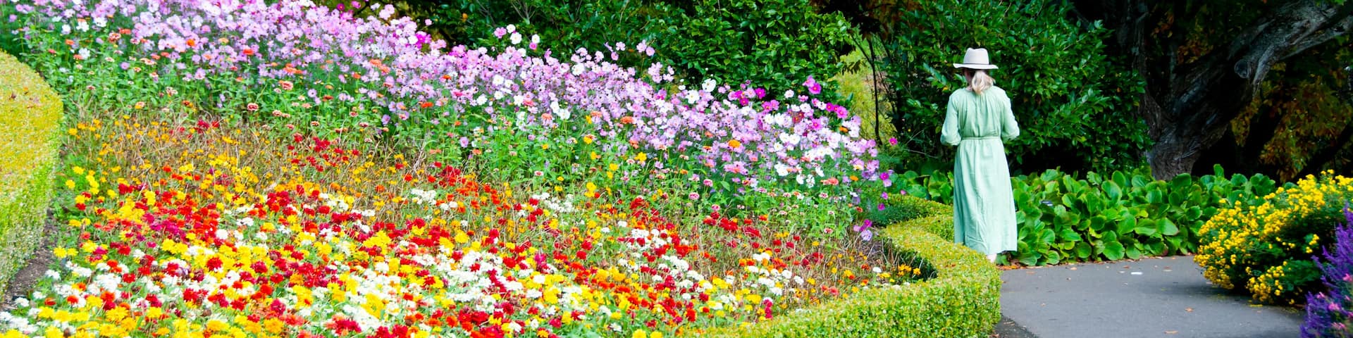 A woman in a green dress and hat walking along a path lined with colorful flowers and trees.