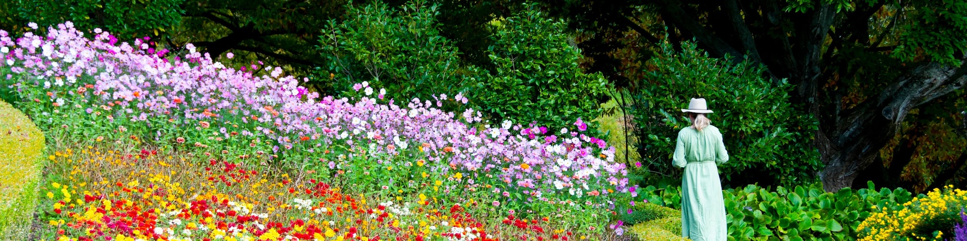 Eine Frau in einem grünen Kleid und Hut geht einen Weg entlang, der von bunten Blumen und Bäumen gesäumt ist.
