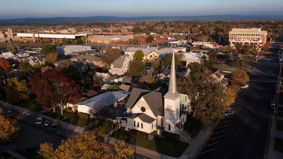 A church with a tall steeple, surrounded by residential houses and a large industrial building in the background.