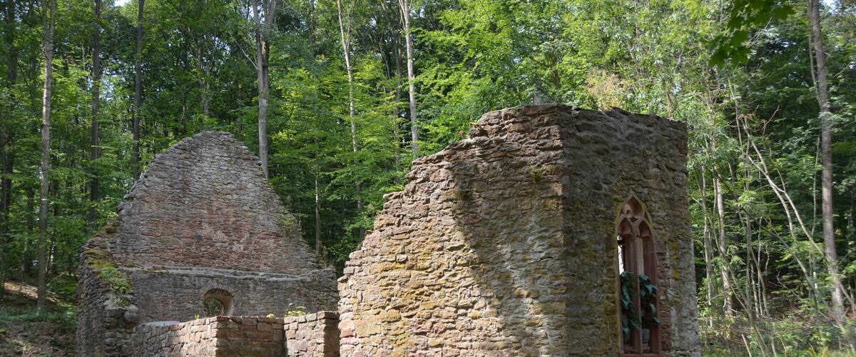 A stone ruin with a small tower and arched window surrounded by a forest.