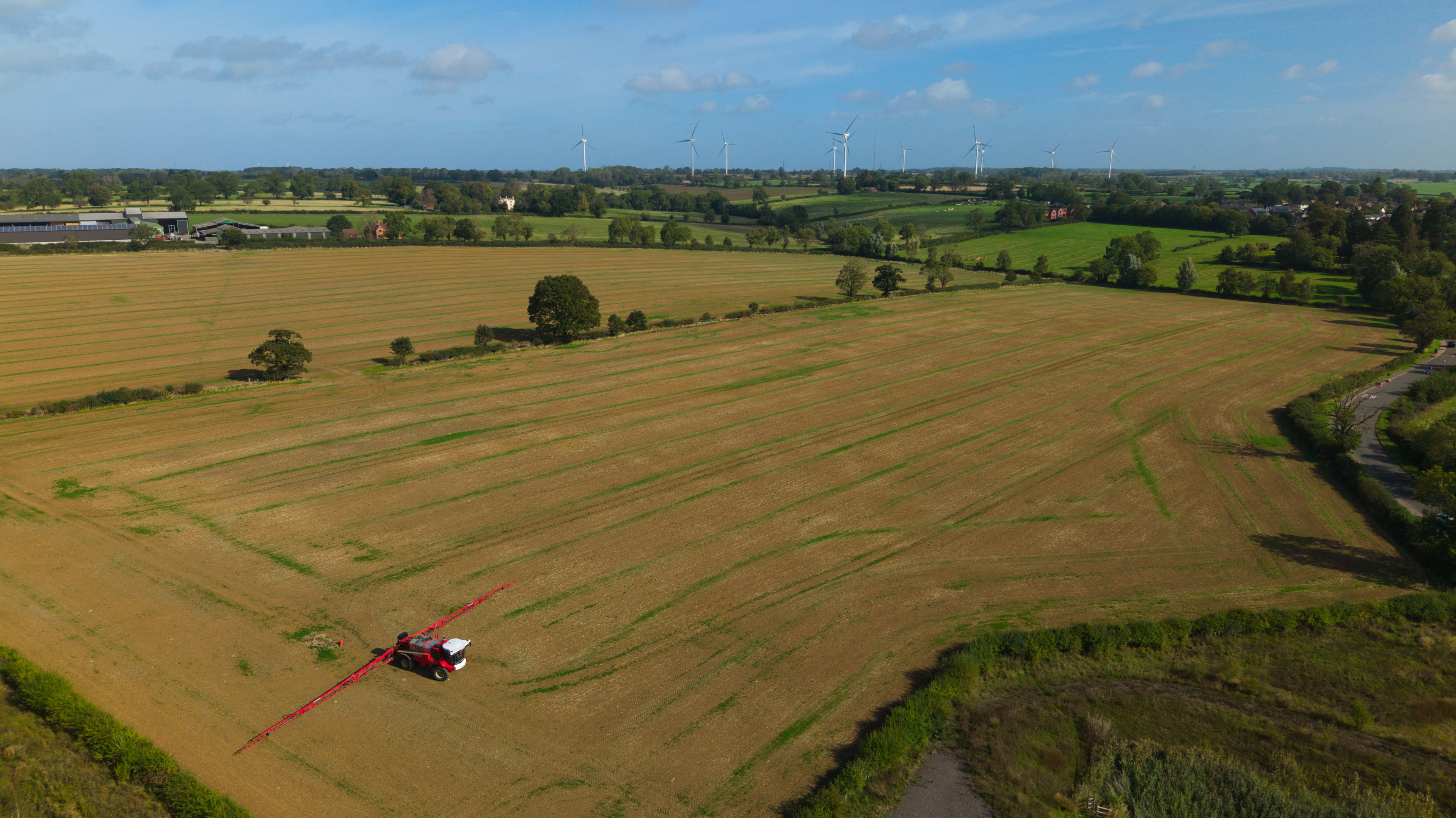 A helicopter flying over a vast agricultural field with wind turbines in the background.