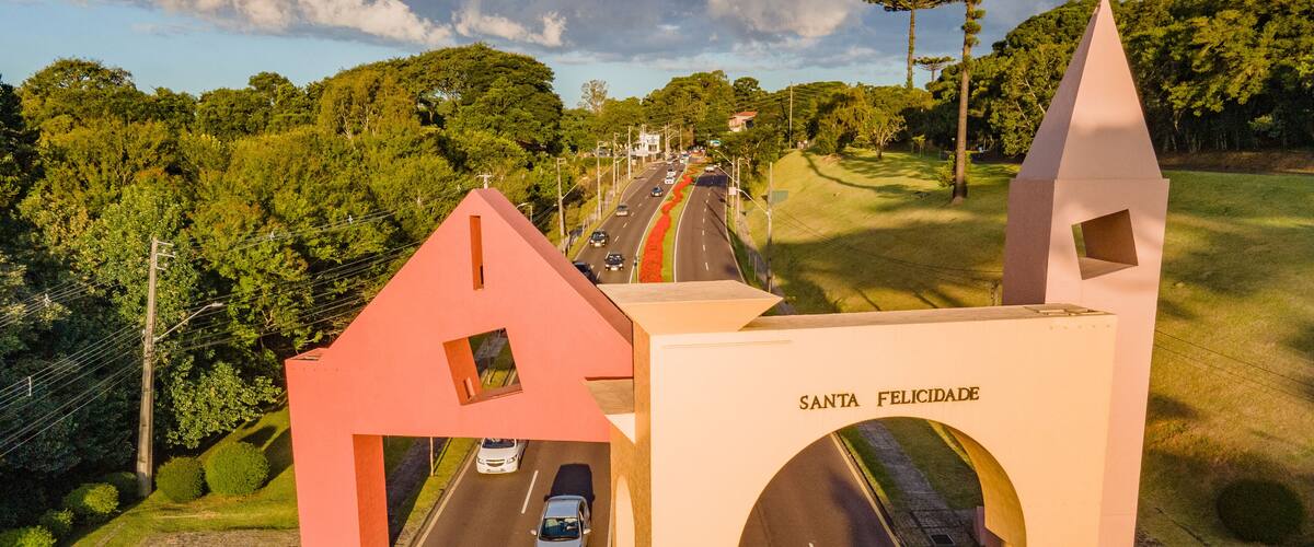 A road passing through a rural area with a large archway labeled "SANTA FELICIDADE" and a distinctive red house-like structure.