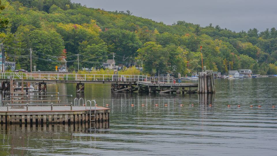 A wooden pier extending into a calm lake, surrounded by lush green hills and a few buildings in the background.