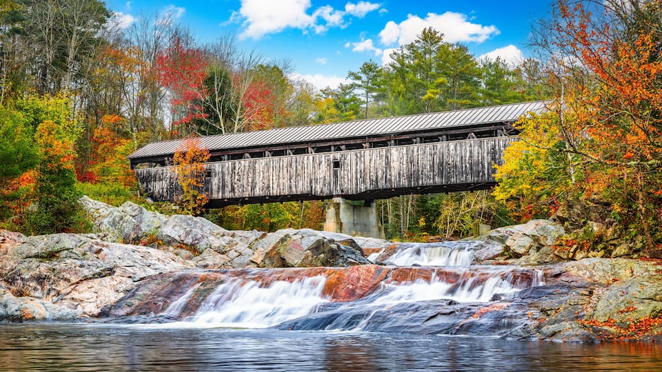 A wooden covered bridge over a waterfall in a forested area with autumn foliage.