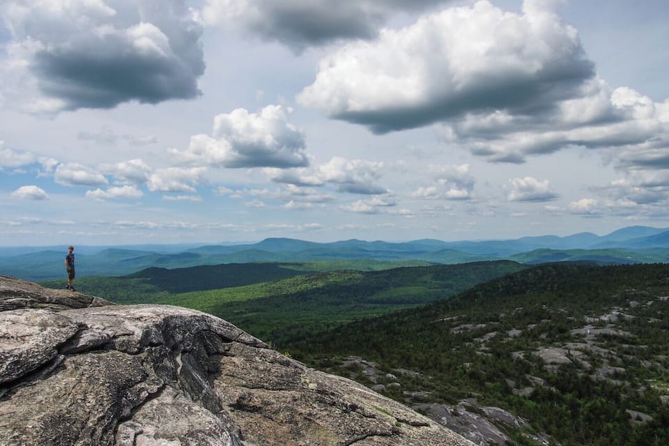 A person standing on a rocky outcrop overlooking a vast mountainous landscape with rolling hills and a partly cloudy sky.
