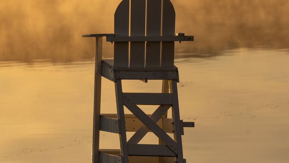 A wooden Adirondack chair on a grassy shore with a misty lake and hills in the background.
