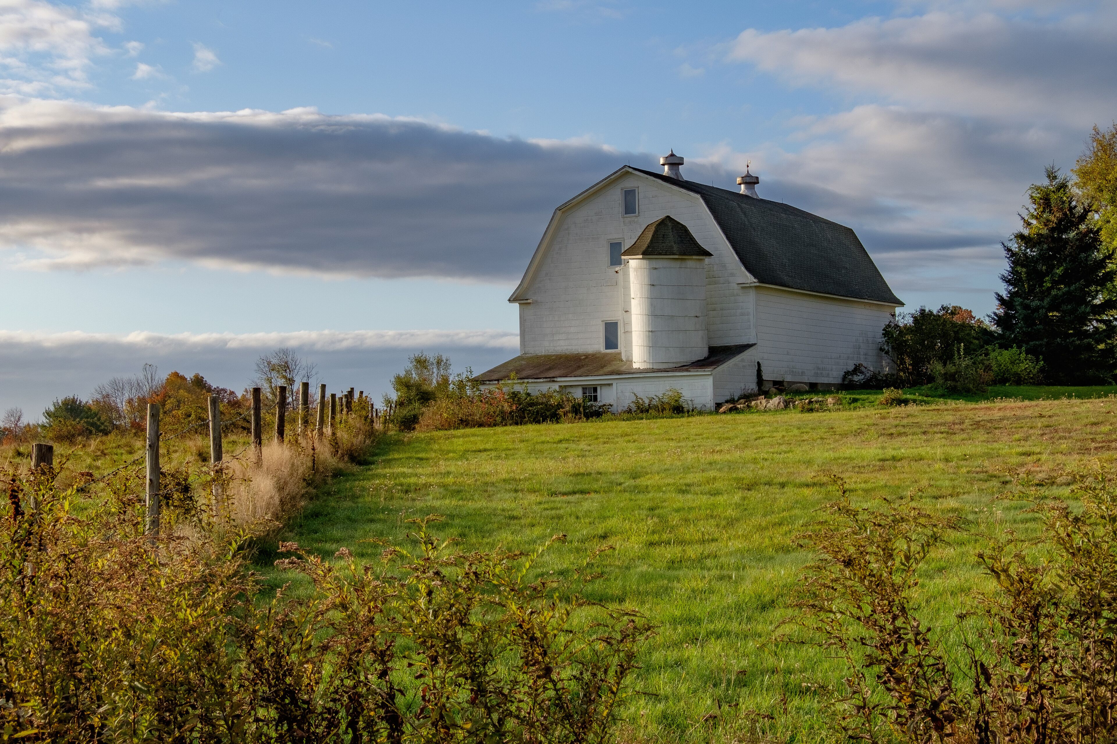 A white barn with a silo on a grassy hill.
