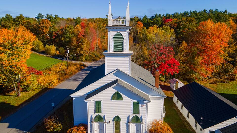 A white church with a steeple and green shutters, surrounded by autumn foliage.