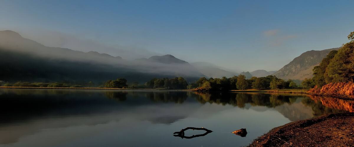 A calm lake with reflections of trees and mountains, surrounded by a forested landscape.
