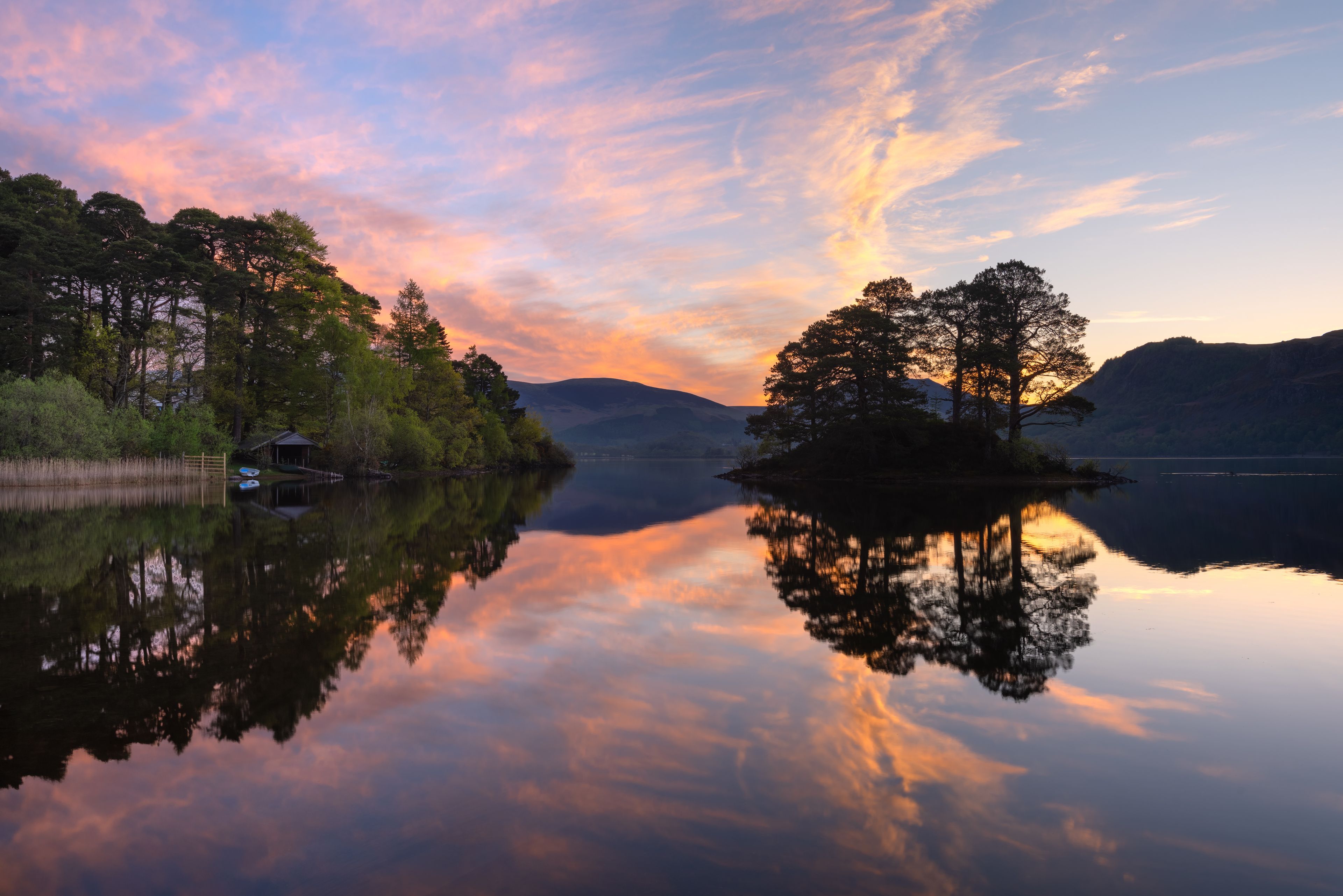Un lago tranquilo con reflejos de árboles y una casa, rodeado de colinas y un cielo colorido.