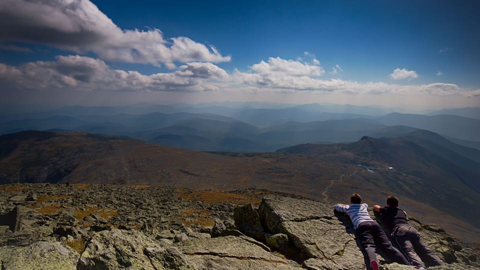 Two people lying on a rocky mountain ridge with a view of distant mountain ranges and a clear sky.