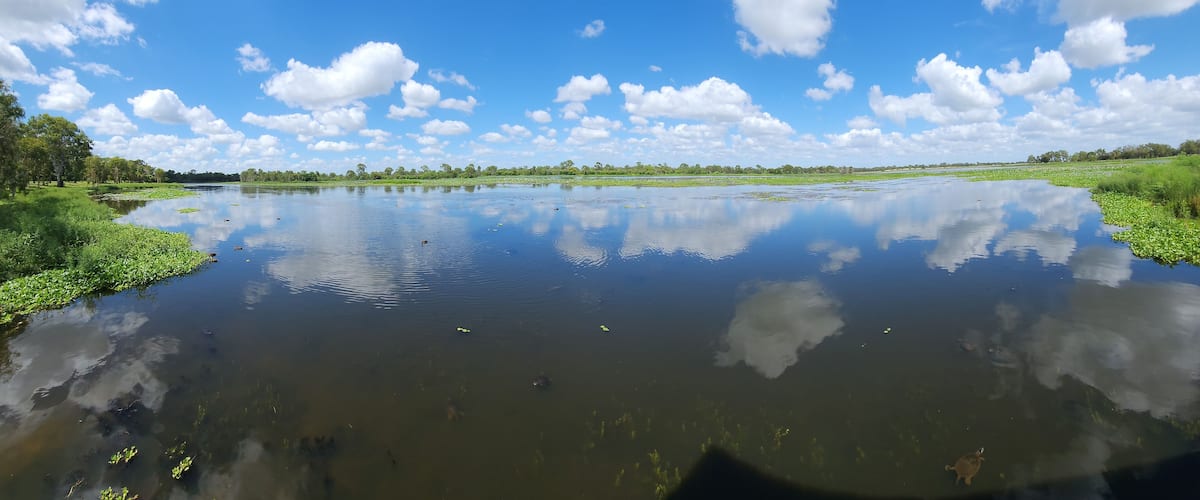A calm lake with reflections of the sky and clouds, surrounded by greenery and trees.