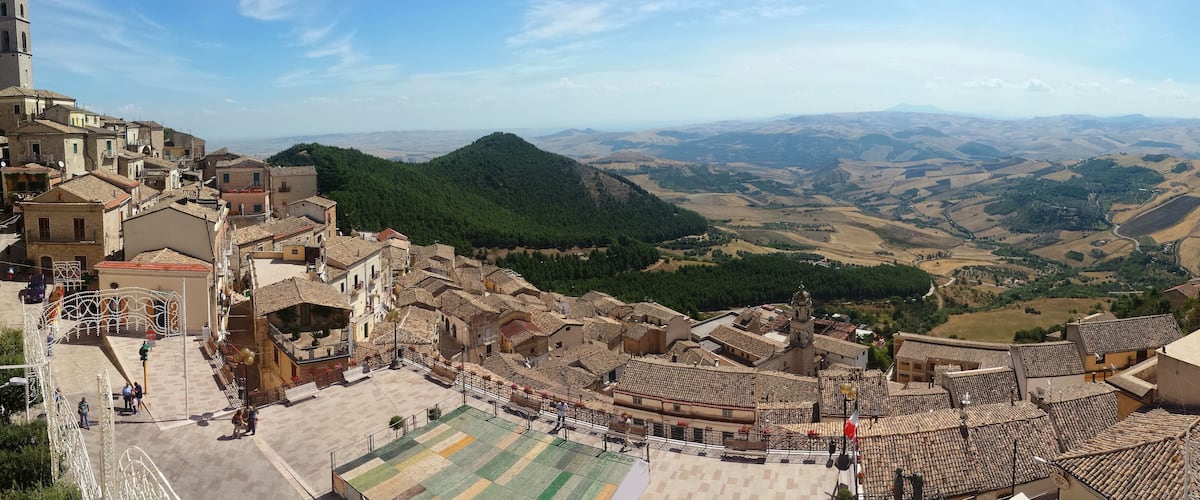 A historic town with terracotta-roofed buildings, a central square, and a mountainous backdrop.