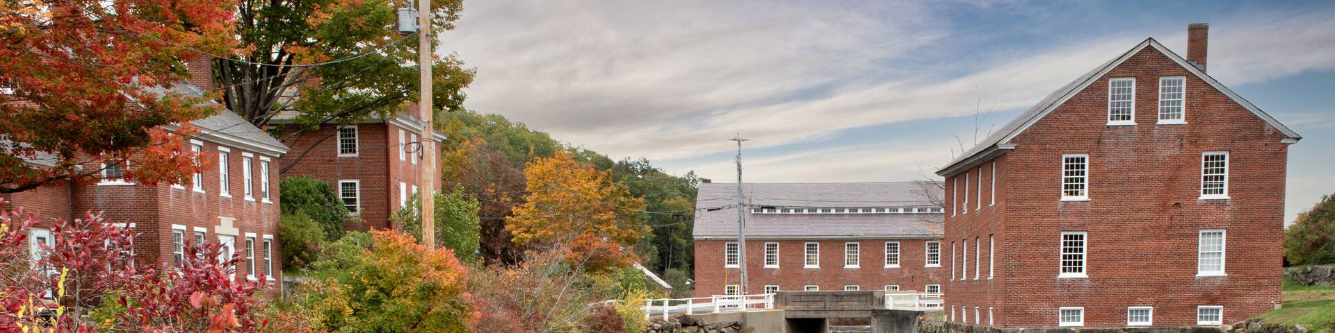 Colorful autumn scene at historic 19th-century textile mill town in Monadnock region of New Hampshire. Picturesque village of Harrisville with reflection of sky and red brick buildings on water.