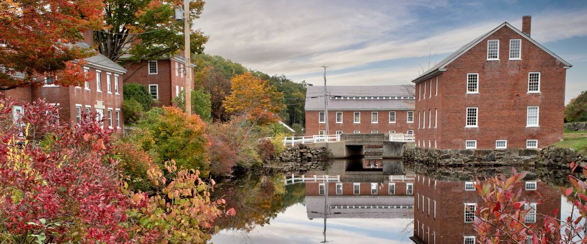 A calm river with a bridge, surrounded by autumn foliage and historic brick buildings.