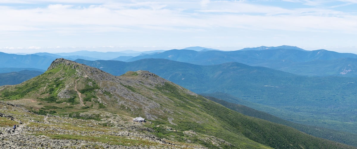 A mountain peak with a small cabin, surrounded by forested slopes and distant mountain ranges under a clear sky.