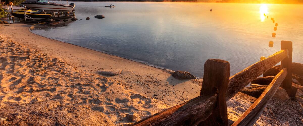 A calm lake at sunset with boats, a wooden dock, and a sandy beach.