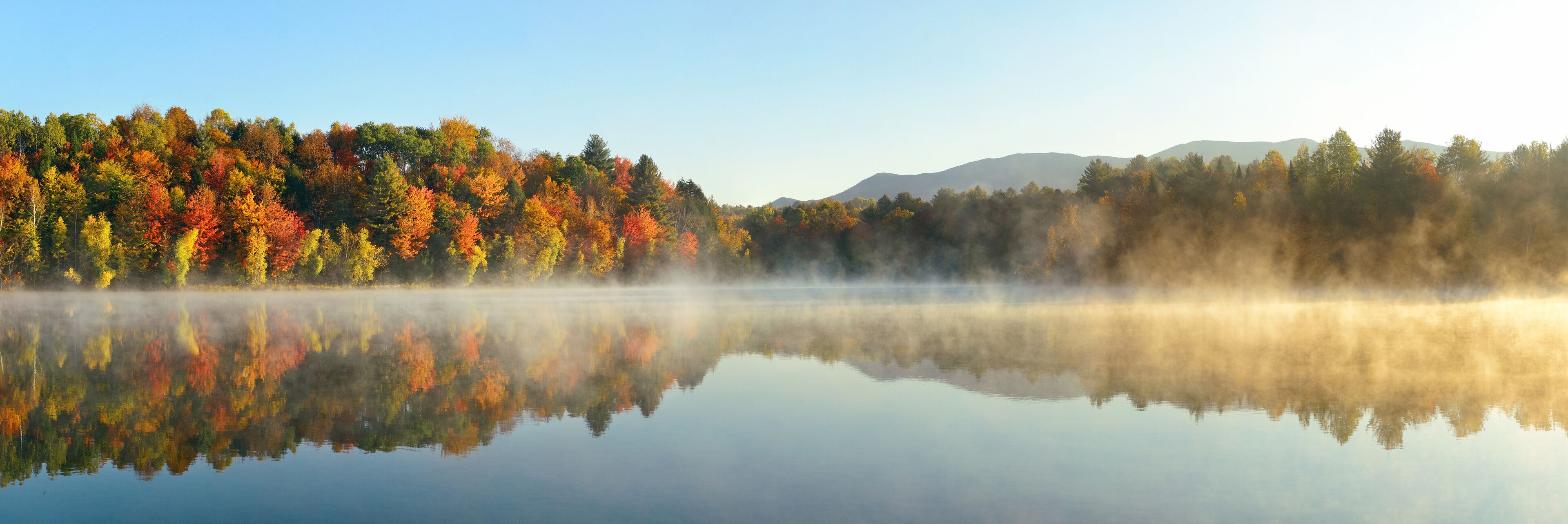 A calm lake reflecting colorful autumn trees and misty mountains in the distance.