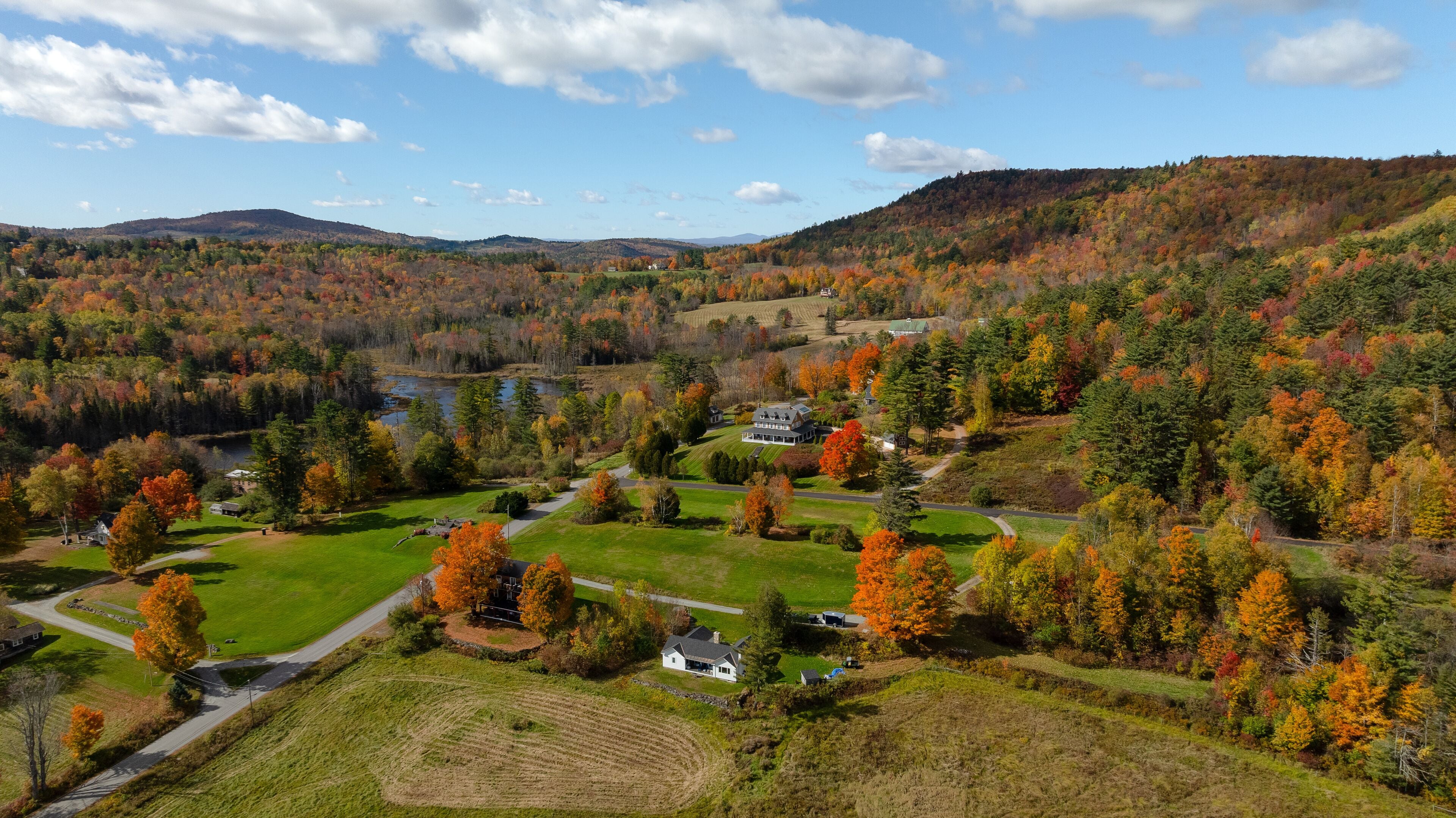 Aerial view of vibrant fall foliage painting the landscape around scattered houses and a tranquil pond, with rolling hills beyond, in the White Mountains. Sugar Hill, New Hampshire, United States.