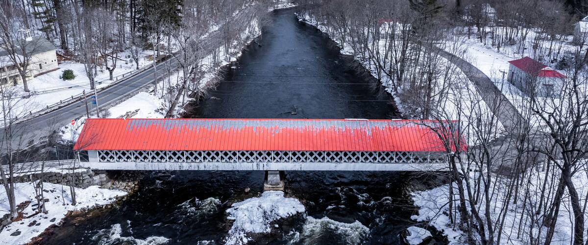 A snow-covered bridge over a river with a red roof, surrounded by trees and buildings.