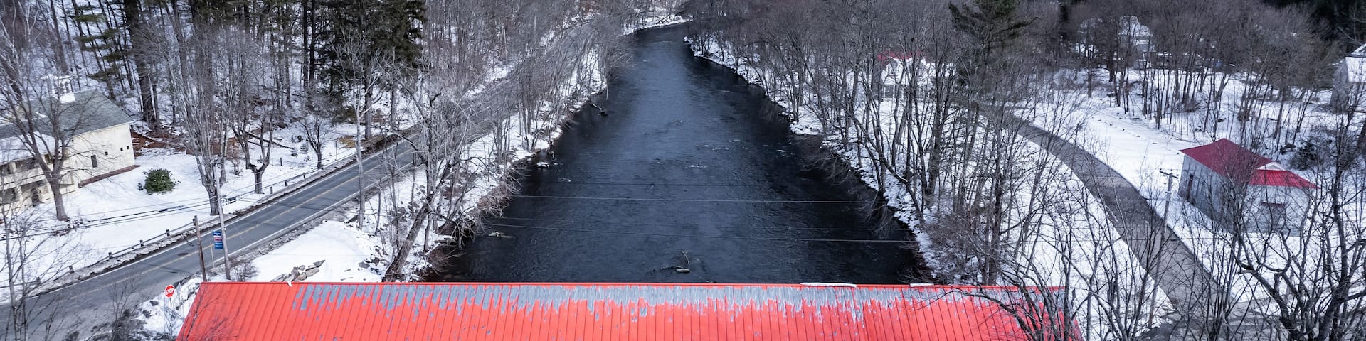 A snow-covered bridge over a river with a red roof, surrounded by trees and buildings.