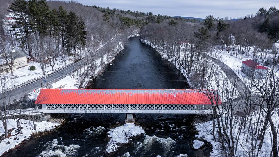 A snow-covered bridge over a river with a red roof, surrounded by trees and buildings.