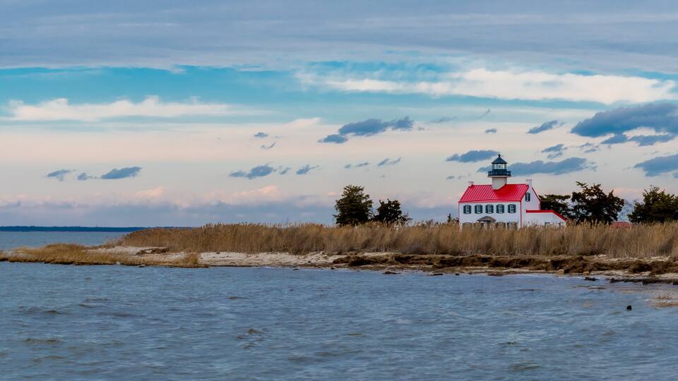 A lighthouse on a sandy spit with tall grasses, a red roof, and a cloudy sky.