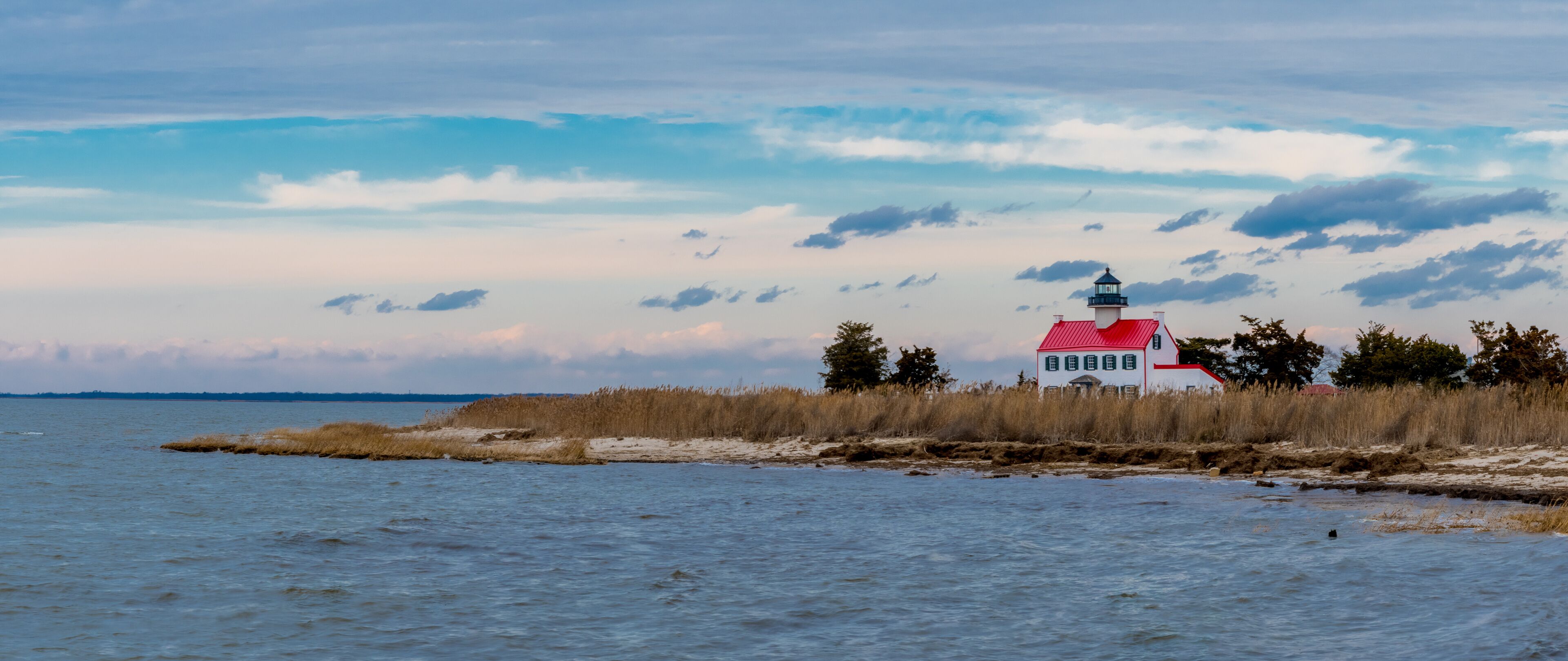 A lighthouse on a sandy spit with tall grasses, a red roof, and a cloudy sky.