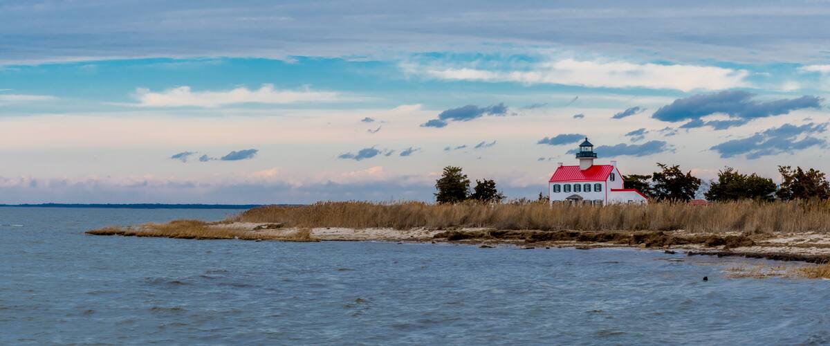A lighthouse on a sandy spit with tall grasses, a red roof, and a cloudy sky.