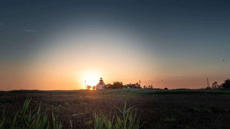 A sunset over a field with a house and trees in the background.