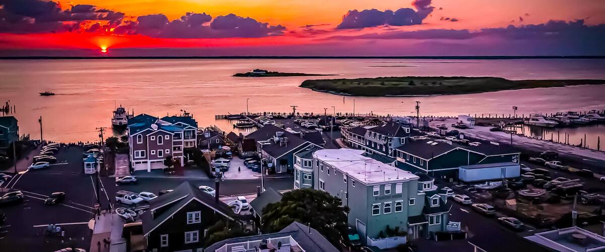 A coastal town at sunset with a marina, boats, and a prominent building with a green roof.