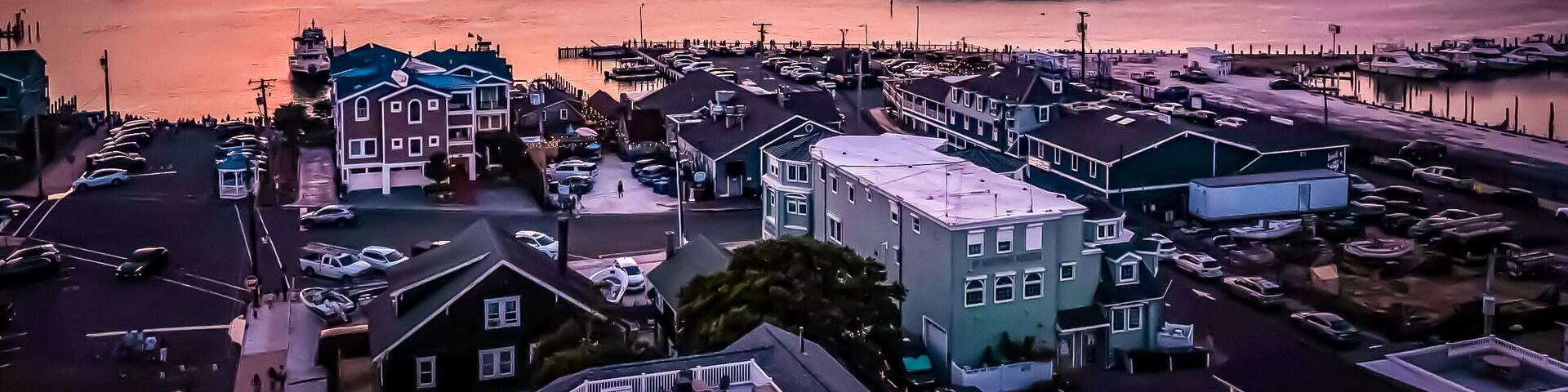 A coastal town at sunset with a marina, boats, and a prominent building with a green roof.