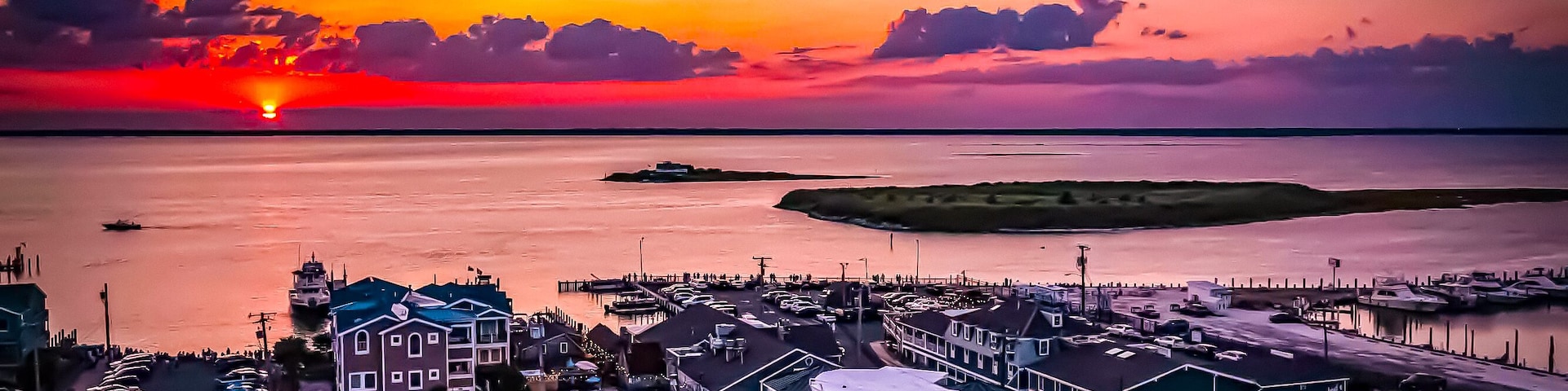 A coastal town at sunset with a marina, boats, and a prominent building with a green roof.