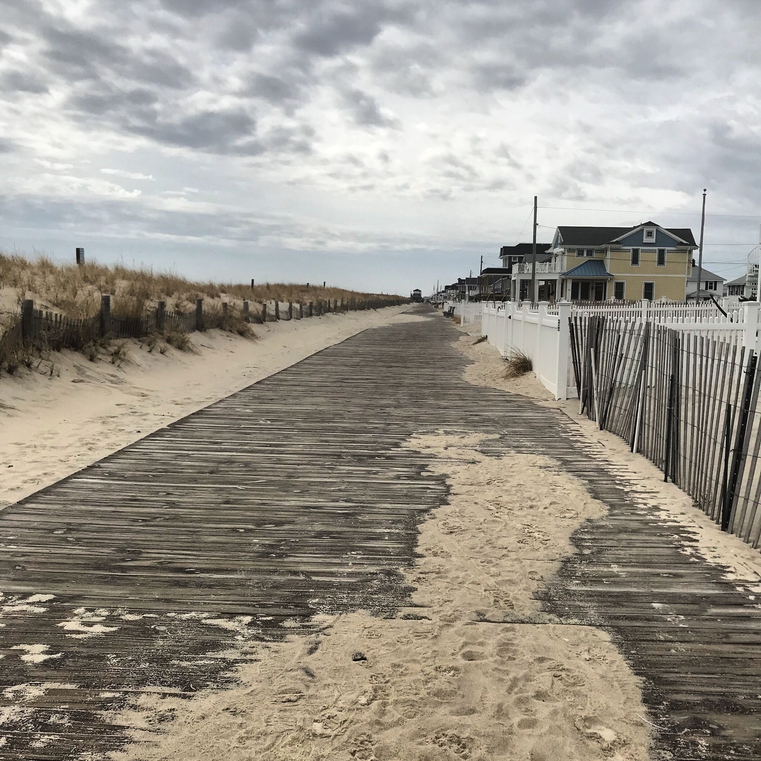 A wooden boardwalk along a sandy beach with a white fence and houses in the background.