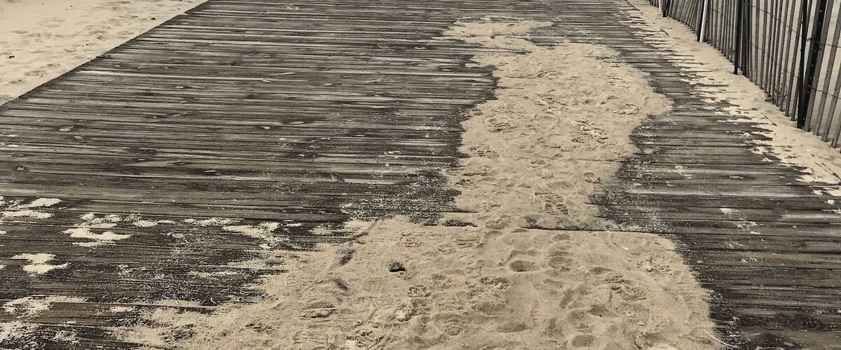 A wooden boardwalk along a sandy beach with a white fence and houses in the background.