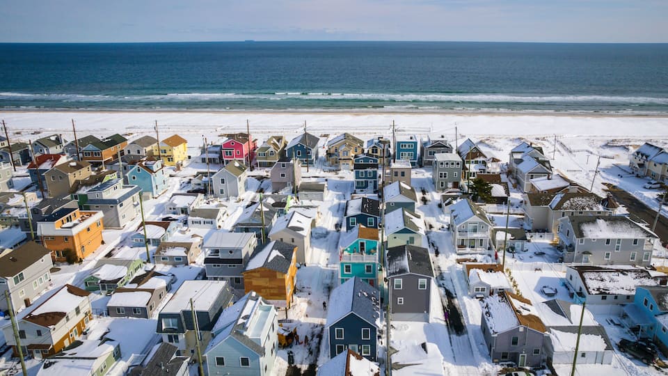 A coastal residential area with houses covered in snow, a beach, and the ocean in the background.