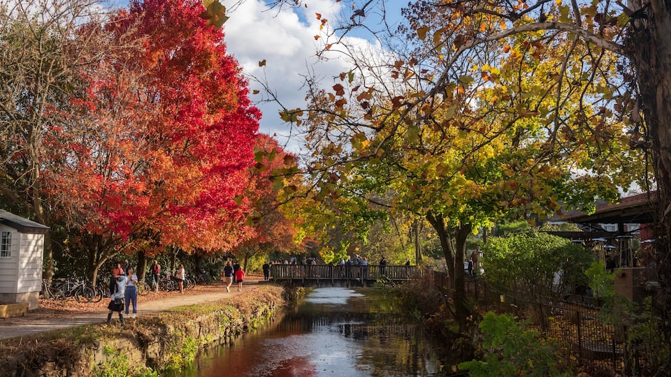 A river with autumn foliage, a wooden bridge, and people walking along a path.