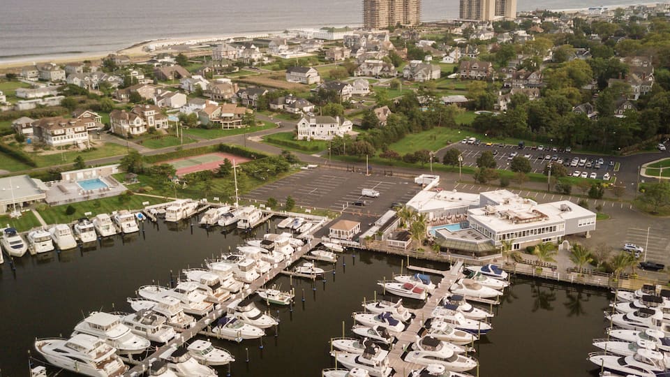 A marina with numerous boats docked, surrounded by residential houses and a beach in the background.