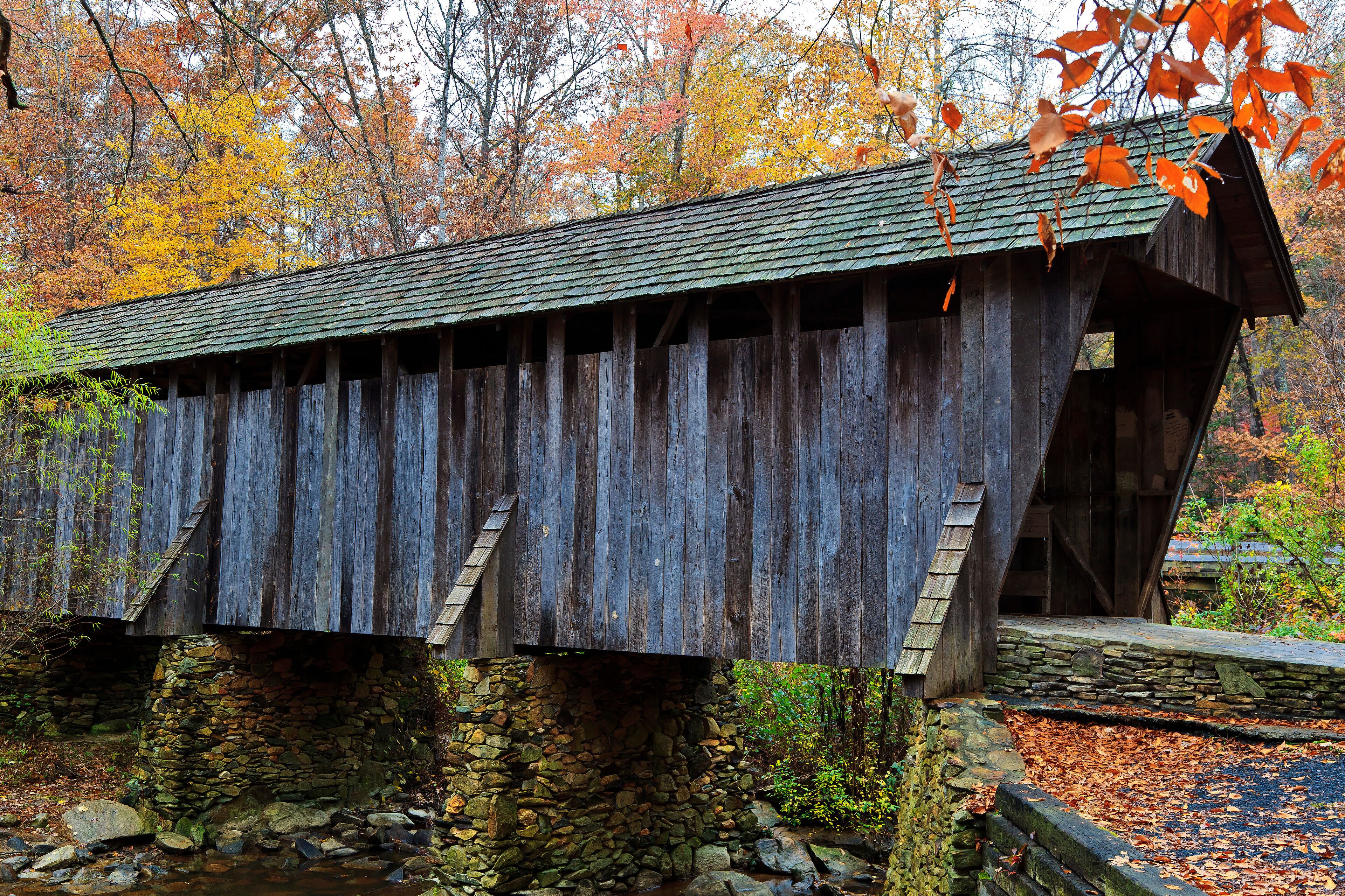 A wooden covered bridge with stone foundations, surrounded by autumn foliage.