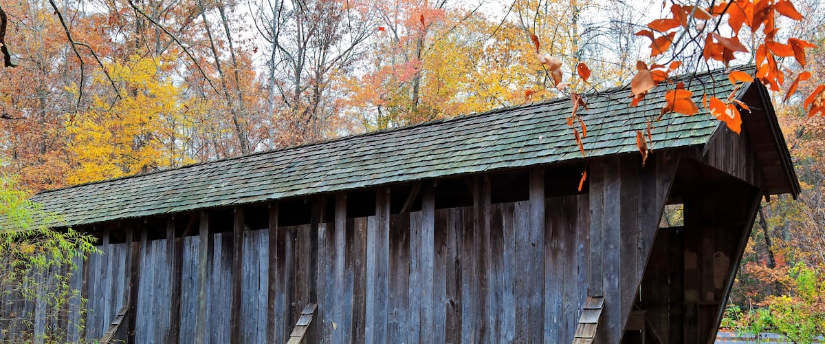 A wooden covered bridge with stone foundations, surrounded by autumn foliage.